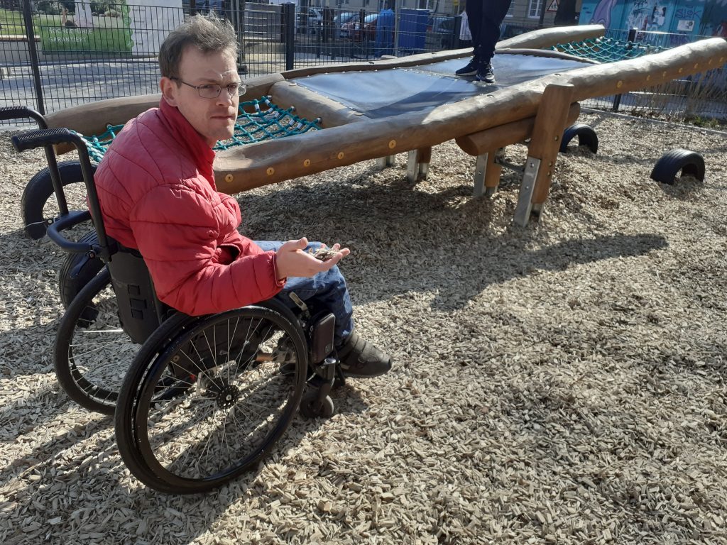 Ein Rollstuhlfahrer steckt mit seinen Rädern im Untergrund eines Spielplatzes fest. Im Hintergrund eine Wippe, die er aber nicht erreichen kann.